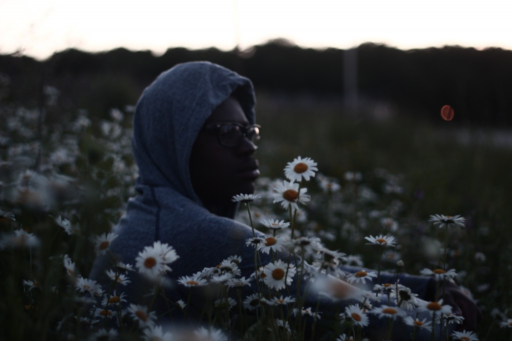 masculine presenting person with dark skin. They are sitting in a field of daisies