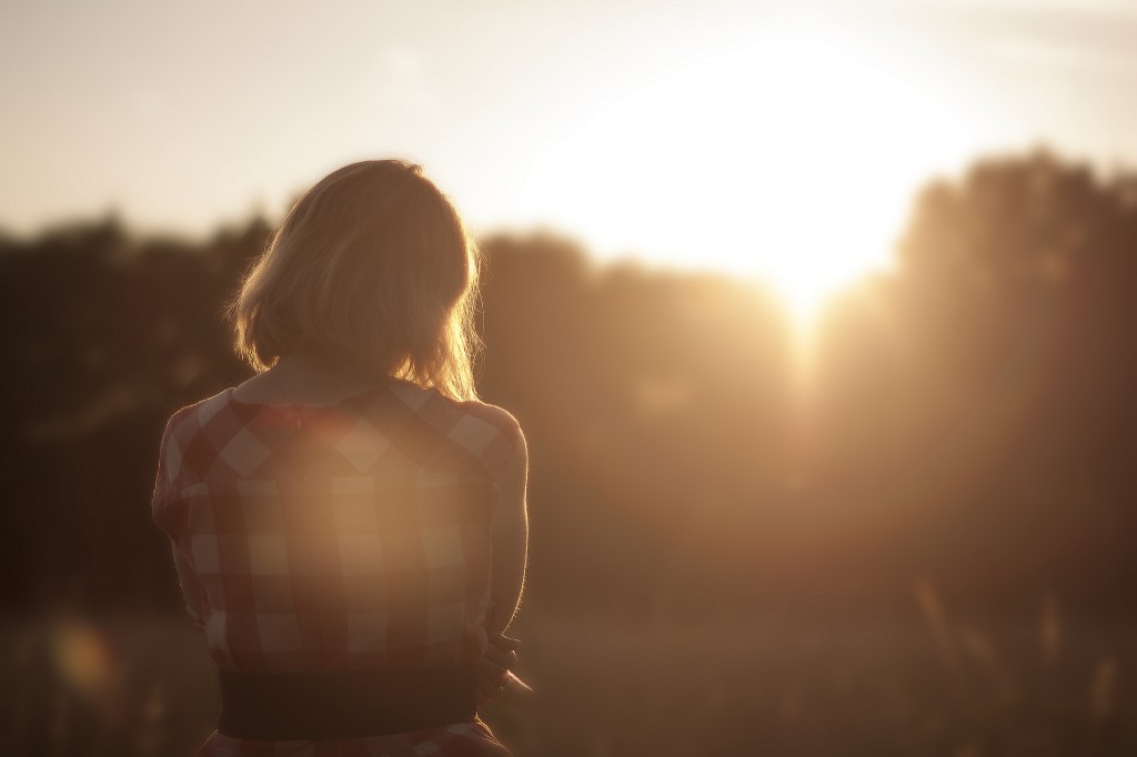 light skin feminine presenting person with blonde hair looking out at the sun in a field. They're wearing a red plaid dress with a black belt around the waist.