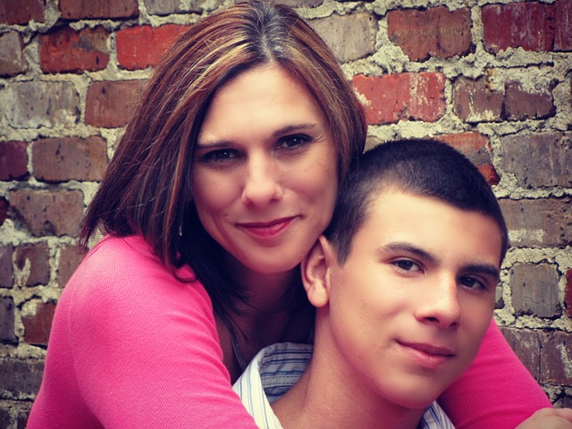mother behind her son hugging him. Her cheek is pressed against his head. They are both smiling. The background is an old red brick wall